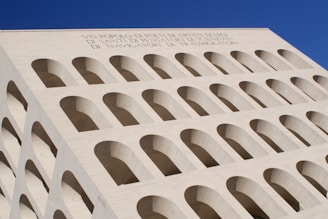 A large, geometrically designed building featuring multiple rows and columns of arches, made from a light-colored stone. The structure has a modernist architectural style with an inscription at the top of the facade. The sky is clear and blue in the background.