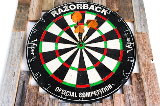 A dartboard is mounted on a wooden surface with three darts successfully hitting close to the bullseye. The dartboard features alternating black and white sections, highlighted with green and red rings. The words 'Razorback' and 'Official Competition' are printed on the board.