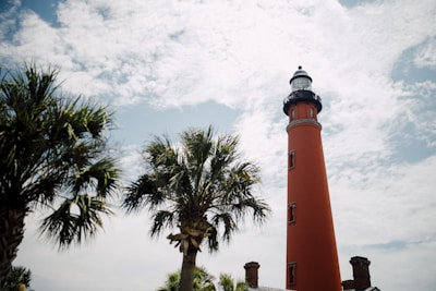 A tall, red lighthouse with a domed top stands prominently against a partly cloudy sky. Several palm trees frame the lighthouse, indicating a coastal or tropical location.