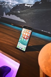 A smartphone with a colorful home screen is propped up against a wooden surface in front of a large computer monitor displaying a landscape with mountains and clouds. The monitor also shows a waveform graphic and some widgets with details like time and date. A computer mouse is visible on a desk with a purple light beneath it, and there is a round object partially visible in the foreground.