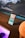 A smartphone with a colorful home screen is propped up against a wooden surface in front of a large computer monitor displaying a landscape with mountains and clouds. The monitor also shows a waveform graphic and some widgets with details like time and date. A computer mouse is visible on a desk with a purple light beneath it, and there is a round object partially visible in the foreground.