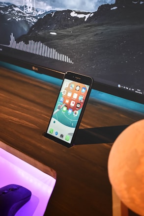 A smartphone with a colorful home screen is propped up against a wooden surface in front of a large computer monitor displaying a landscape with mountains and clouds. The monitor also shows a waveform graphic and some widgets with details like time and date. A computer mouse is visible on a desk with a purple light beneath it, and there is a round object partially visible in the foreground.
