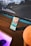 A smartphone with a colorful home screen is propped up against a wooden surface in front of a large computer monitor displaying a landscape with mountains and clouds. The monitor also shows a waveform graphic and some widgets with details like time and date. A computer mouse is visible on a desk with a purple light beneath it, and there is a round object partially visible in the foreground.