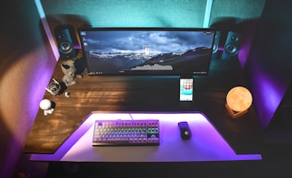 A modern computer setup featuring a wide monitor displaying a scenic mountain wallpaper. The desk is lit with ambient purple and blue lighting. Accessories include a keyboard with colorful backlighting, a mouse, two small figurines, a smartphone, and a spherical lamp resembling the moon.