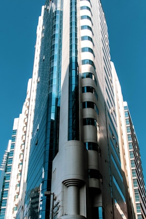 A modern, high-rise skyscraper with sleek architectural design features glass and concrete materials. The building is illuminated by natural sunlight, creating contrasting shadows and highlights.