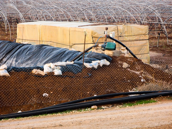 A farming landscape featuring a large plastic-covered structure with a network of metal arches, possibly greenhouses, in the background. A fenced off area includes a piled dirt embankment and a yellow storage container. A pump system is attached, with pipes extending across the scene. Sandbags and black plastic sheeting are used for reinforcement and covering.