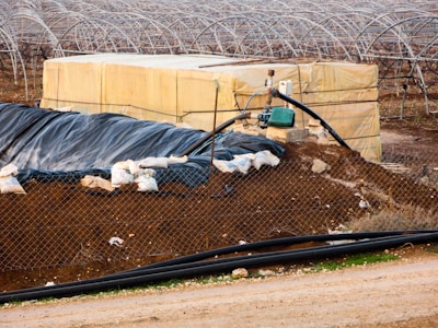 A farming landscape featuring a large plastic-covered structure with a network of metal arches, possibly greenhouses, in the background. A fenced off area includes a piled dirt embankment and a yellow storage container. A pump system is attached, with pipes extending across the scene. Sandbags and black plastic sheeting are used for reinforcement and covering.