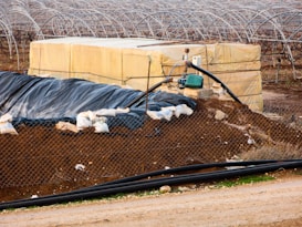 A farming landscape featuring a large plastic-covered structure with a network of metal arches, possibly greenhouses, in the background. A fenced off area includes a piled dirt embankment and a yellow storage container. A pump system is attached, with pipes extending across the scene. Sandbags and black plastic sheeting are used for reinforcement and covering.