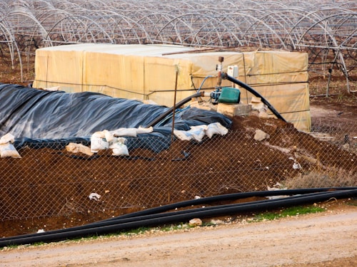 A farming landscape featuring a large plastic-covered structure with a network of metal arches, possibly greenhouses, in the background. A fenced off area includes a piled dirt embankment and a yellow storage container. A pump system is attached, with pipes extending across the scene. Sandbags and black plastic sheeting are used for reinforcement and covering.