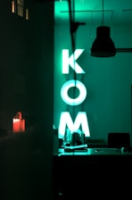 A dimly lit interior space with a neon green sign displaying the letters K, O, and M. The sign is situated in what appears to be an office or workspace setting, with various items on a desk below it. A dark hanging lamp can be seen above the space, and the overall vibe is modern and minimalist. A faint reflection is visible on the left, showing part of a night scene outside.