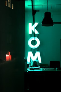 A dimly lit interior space with a neon green sign displaying the letters K, O, and M. The sign is situated in what appears to be an office or workspace setting, with various items on a desk below it. A dark hanging lamp can be seen above the space, and the overall vibe is modern and minimalist. A faint reflection is visible on the left, showing part of a night scene outside.