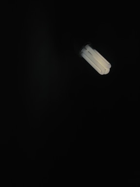 A fluorescent light bulb emits a soft glow in a dark environment, creating a stark contrast between the bright light and the surrounding darkness.