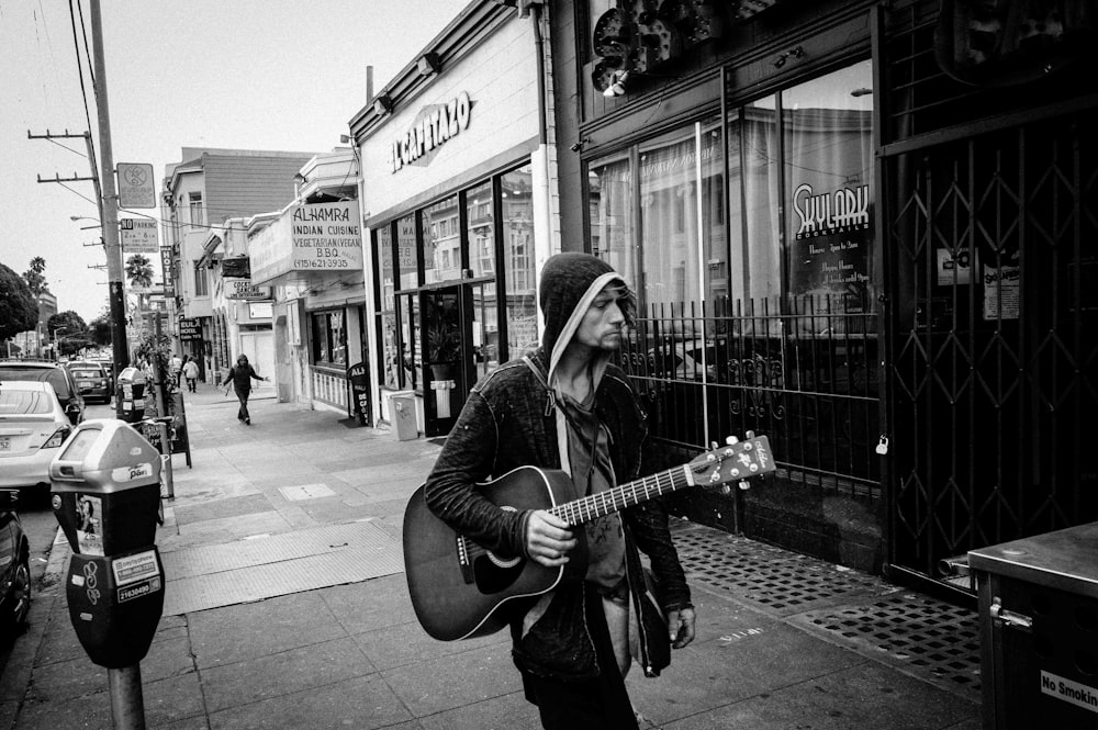 man in hoodie carrying guitar in greyscale photography