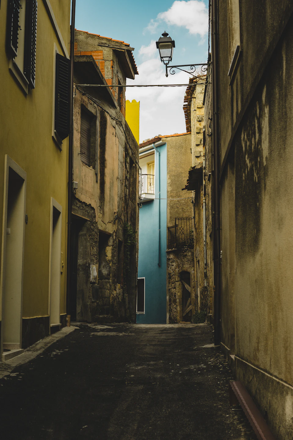 The image depicts a narrow alleyway between two buildings, one painted yellow and the other blue. The alley is flanked by stone walls on both sides, adding an old-world charm to the scene. A white door stands out against the stone wall on the right side of the alley. On the left side, there are three windows with shutters, providing a glimpse into the interior of one of the buildings. Above the alleyway, a street lamp hangs from a wire, casting light onto the cobblestone path below and illuminating the scene.
