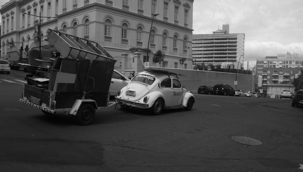 A compact utility trailer hooked up to a passenger car on a sunny city street.