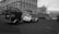 A vintage Volkswagen Beetle is towing a small, overloaded trailer on an empty city street. The scene features a large, classical-style building with arched windows on a cloudy day. Several modern buildings are visible in the background, adding contrast to the historical architecture.