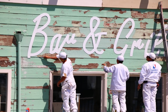 Three workers are painting the exterior of a building with a weathered wooden facade in a light mint green color. They are wearing white uniforms with logos on the back. The building has large white script letters spelling out 'Bar & Grill.' A ladder is leaning against the wall, and the sign appears to be in the process of being refurbished.
