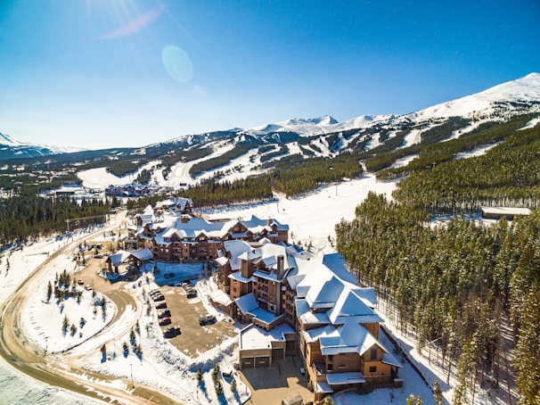 A snowy mountain resort with a clear blue sky, surrounded by expansive forests. Several buildings with snow-covered roofs are visible, likely accommodations or lodges. In the background, ski slopes stretch across the landscape, indicating a ski resort area. A winding road leads to the lodges, and a parking lot is partially filled with vehicles.