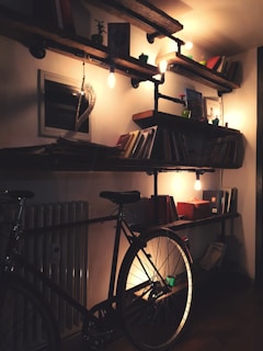 A cozy interior with wooden shelves on a wall, filled with books and small decor items. Warm lighting from exposed bulbs creates a soft glow. A bicycle is leaning against the wall beneath the shelves.