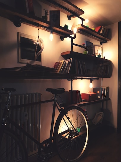 A cozy interior with wooden shelves on a wall, filled with books and small decor items. Warm lighting from exposed bulbs creates a soft glow. A bicycle is leaning against the wall beneath the shelves.