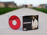 A CD case and disc are positioned on a cement walkway. The CD features a bold red color with the name 'EMINEM' in black letters. The case displays a black and white image of a man in a puffy coat with the text 'KILL YOU' in red. The background is a blurred suburban street.