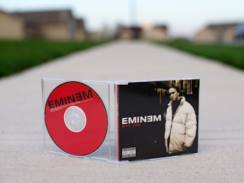 A CD case and disc are positioned on a cement walkway. The CD features a bold red color with the name 'EMINEM' in black letters. The case displays a black and white image of a man in a puffy coat with the text 'KILL YOU' in red. The background is a blurred suburban street.