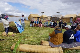 A group of people is seated on a reed-covered ground, listening to a local presenter. In the background, traditional reed huts and solar panels can be seen, with a large lake and mountains further away under a cloudy sky.