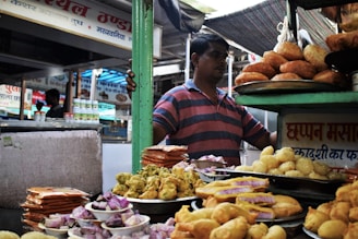 A food stall at a market is displaying a variety of Indian snacks. Plates are piled high with items like fried pastries, puris, and colorful sweets. A person is standing behind the stall, wearing a striped shirt and surrounded by signs and various containers.