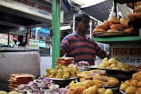 A food stall at a market is displaying a variety of Indian snacks. Plates are piled high with items like fried pastries, puris, and colorful sweets. A person is standing behind the stall, wearing a striped shirt and surrounded by signs and various containers.