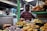 A food stall at a market is displaying a variety of Indian snacks. Plates are piled high with items like fried pastries, puris, and colorful sweets. A person is standing behind the stall, wearing a striped shirt and surrounded by signs and various containers.