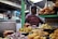 A food stall at a market is displaying a variety of Indian snacks. Plates are piled high with items like fried pastries, puris, and colorful sweets. A person is standing behind the stall, wearing a striped shirt and surrounded by signs and various containers.