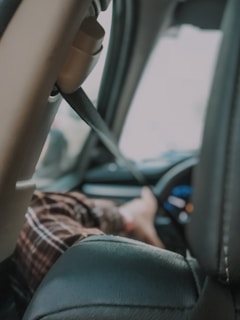 A view inside a car showing a pair of feet resting on the dashboard. The car interior includes visible elements like a seatbelt and steering wheel. The person appears relaxed with their legs on a headrest and visible plaid pajamas.
