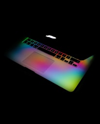 A laptop keyboard is illuminated with rainbow colors against a dark background. The colors create a vibrant gradient over the keyboard surface.