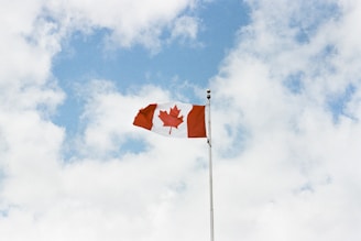 A Canadian flag with a red maple leaf in the center flutters against a backdrop of a partly cloudy sky.