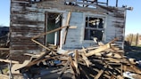 A partially collapsed and abandoned wooden house with peeling paint and exposed beams. Debris and wood planks are scattered in front of the structure, and the interior is visible through broken windows and doorways.
