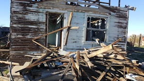 A partially collapsed and abandoned wooden house with peeling paint and exposed beams. Debris and wood planks are scattered in front of the structure, and the interior is visible through broken windows and doorways.