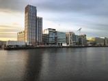 A modern urban landscape featuring a line of contemporary buildings along a waterfront. The tallest structure has a prominent glass facade and the words 'CAPITAL DOCK'. There's a construction crane on one of the buildings, indicating ongoing development. The sky is overcast with a hint of sunlight, reflecting soft colors on the water.
