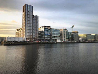 A modern urban landscape featuring a line of contemporary buildings along a waterfront. The tallest structure has a prominent glass facade and the words 'CAPITAL DOCK'. There's a construction crane on one of the buildings, indicating ongoing development. The sky is overcast with a hint of sunlight, reflecting soft colors on the water.