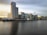 A modern urban landscape featuring a line of contemporary buildings along a waterfront. The tallest structure has a prominent glass facade and the words 'CAPITAL DOCK'. There's a construction crane on one of the buildings, indicating ongoing development. The sky is overcast with a hint of sunlight, reflecting soft colors on the water.