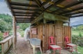 A rustic outdoor structure with a wooden shed that has a corrugated metal roof. Two red chairs are placed in front of the shed, next to a metal washbasin on a table. The setting includes greenery and a small greenhouse tunnel, with a background of lush trees and hills.