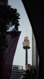 A tall communication tower with a spherical observation deck is surrounded by modern high-rise buildings. There is some greenery and an advertising banner in the foreground, adding depth to the urban scene.
