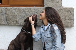 A young woman with long, wavy dark hair wearing a denim jacket is petting and smiling at a brown dog. The dog appears content as it is being touched under its chin. They are outside, next to a stone and white plaster wall with a wooden window frame above them.