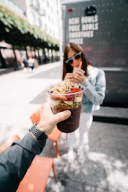 A hand holding a colorful acai bowl topped with sliced bananas, strawberries, blueberries, and granola. In the background, a person is sipping from a smoothie with a straw, wearing sunglasses and a light denim jacket. The setting appears to be outdoors near a food truck offering acai bowls, poke bowls, and smoothies.