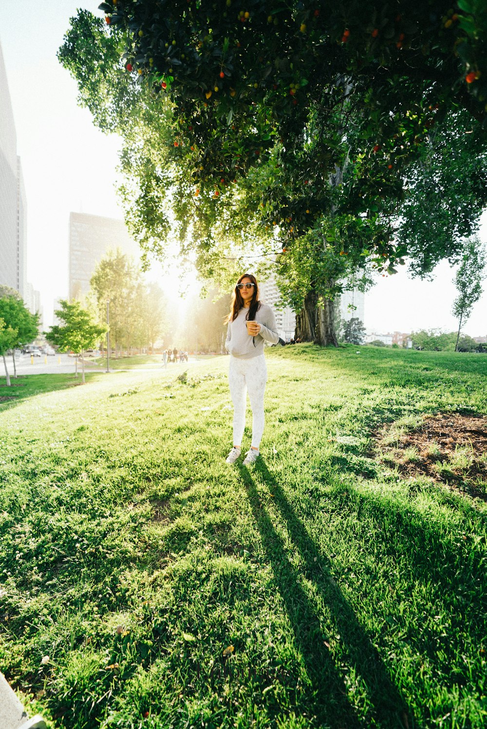woman in white pants and grey sweater stands under green tree