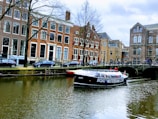 A canal with a tour boat passing under a bridge in a historic European city. The surrounding buildings are characterized by tall windows and traditional architecture. Cars are parked along the street, and a few people can be seen on the boat and by the canal.