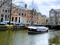 A canal with a tour boat passing under a bridge in a historic European city. The surrounding buildings are characterized by tall windows and traditional architecture. Cars are parked along the street, and a few people can be seen on the boat and by the canal.