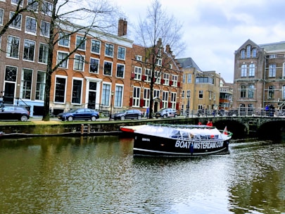 A canal with a tour boat passing under a bridge in a historic European city. The surrounding buildings are characterized by tall windows and traditional architecture. Cars are parked along the street, and a few people can be seen on the boat and by the canal.