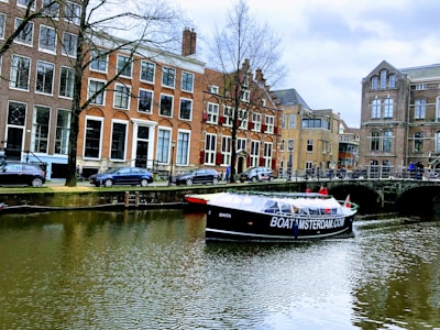 A canal with a tour boat passing under a bridge in a historic European city. The surrounding buildings are characterized by tall windows and traditional architecture. Cars are parked along the street, and a few people can be seen on the boat and by the canal.