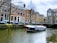 A canal with a tour boat passing under a bridge in a historic European city. The surrounding buildings are characterized by tall windows and traditional architecture. Cars are parked along the street, and a few people can be seen on the boat and by the canal.