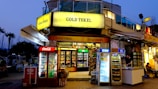 A brightly lit corner store named 'Gold Tekel' located on a busy street, featuring multiple refrigerators filled with beverages from brands like Coca-Cola, Pepsi, and Erikli. A vendor in the adjacent kiosk is serving a customer. There are visible advertisements and branding from Algida and other products. In the background, a few palm trees can be seen against a dusky sky.